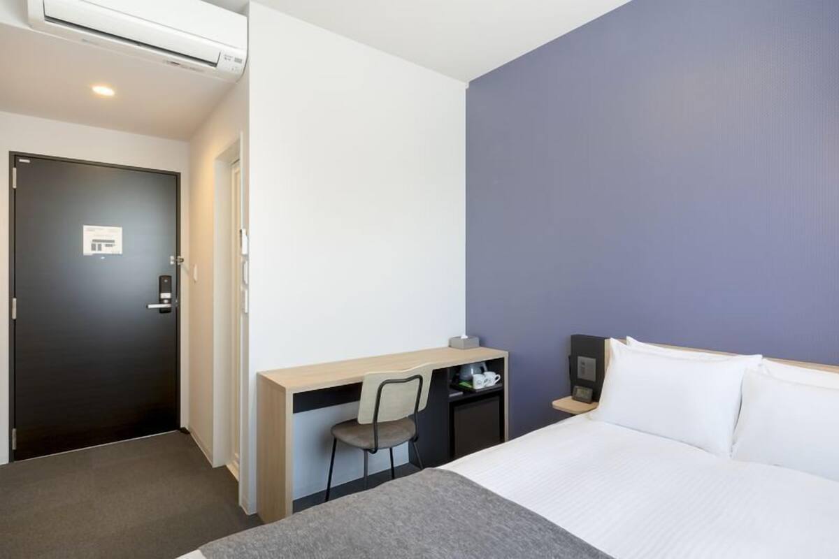 Quarto de hotel com cama, escrivaninha, cadeira, mesa de cabeceira e luminária fixada na parede.