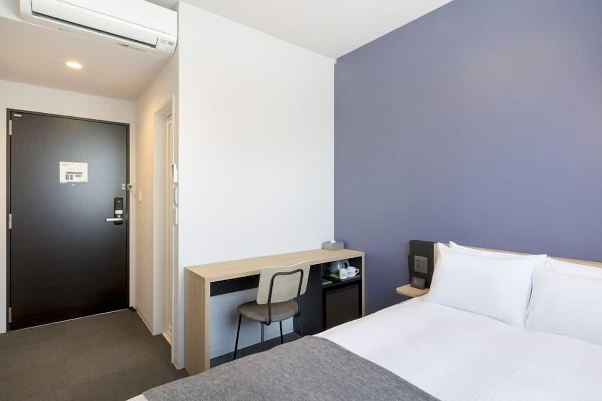 A hotel room with a bed, a desk, a chair, a nightstand, and a wall-mounted lamp.
