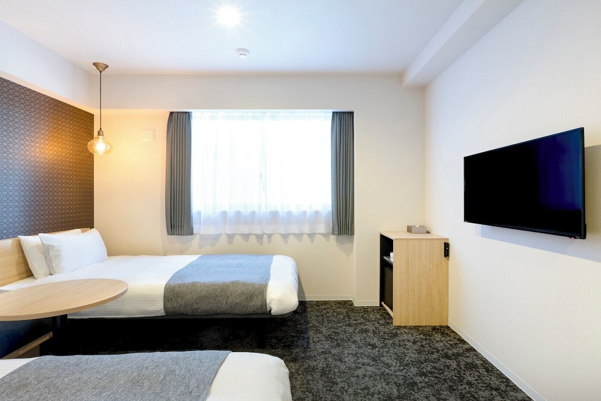 Quarto de hotel com cama, mesa de cabeceira, televisão e janela com cortinas.