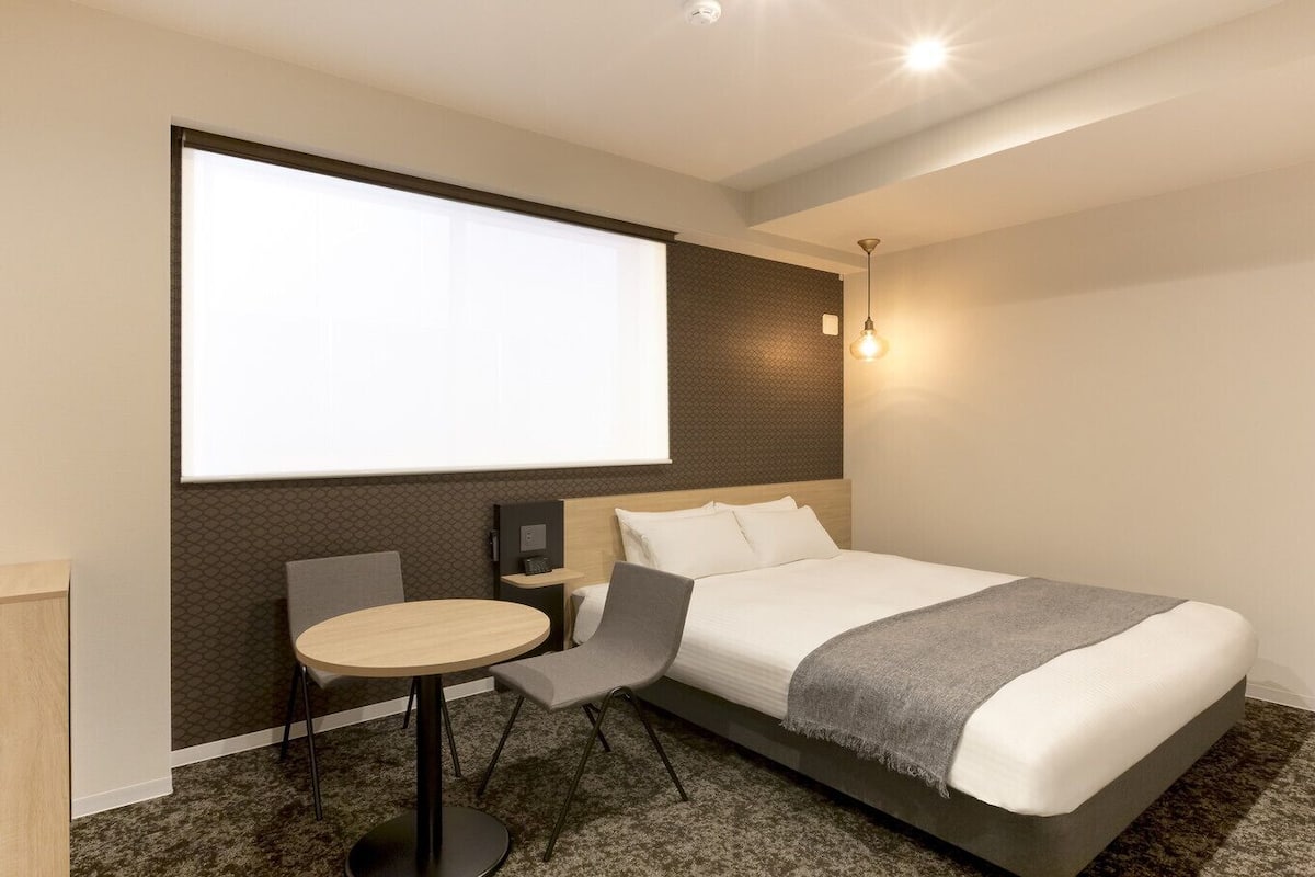 Quarto de hotel com cama, escrivaninha, cadeira, tela de projetor e luminária fixada na parede.