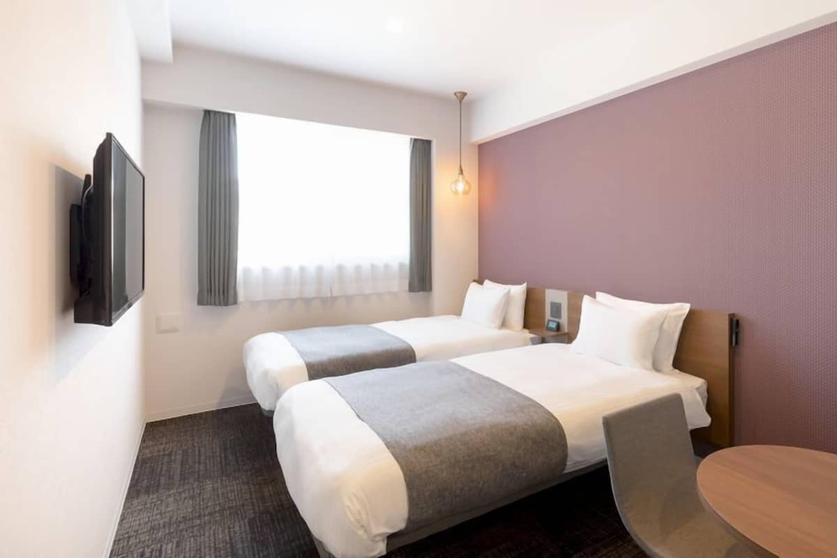 Quarto de hotel com duas camas, televisão, janela com cortinas, pequena mesa e luminária fixada na parede.
