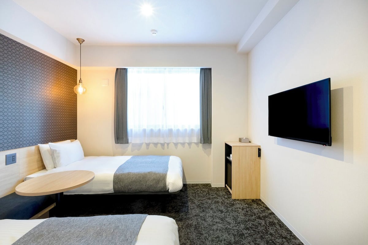 Quarto de hotel com uma cama, uma escrivaninha, uma televisão e uma janela com cortinas.