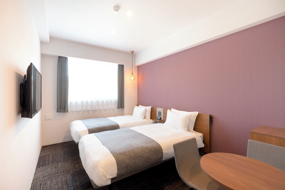 Quarto de hotel com duas camas, televisão, escrivaninha e janela com cortinas.