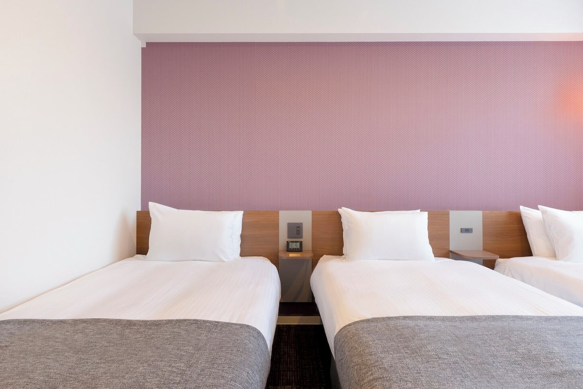 Quarto de hotel com duas camas de solteiro, cabeceira de madeira, mesa de cabeceira com relógio e parede texturizada em tom de rosa.