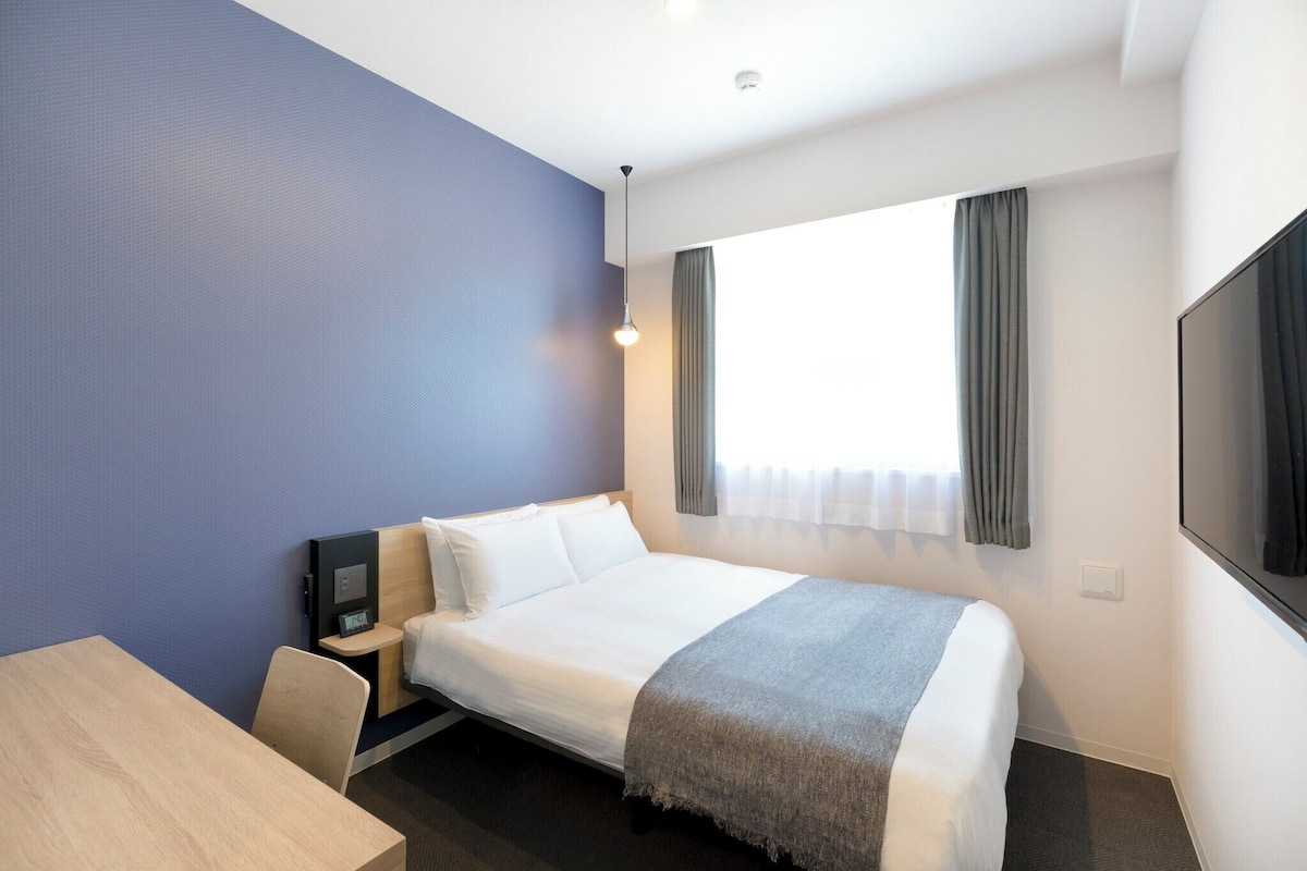 Quarto de hotel com cama, escrivaninha, cadeira, televisão e janela com cortinas.