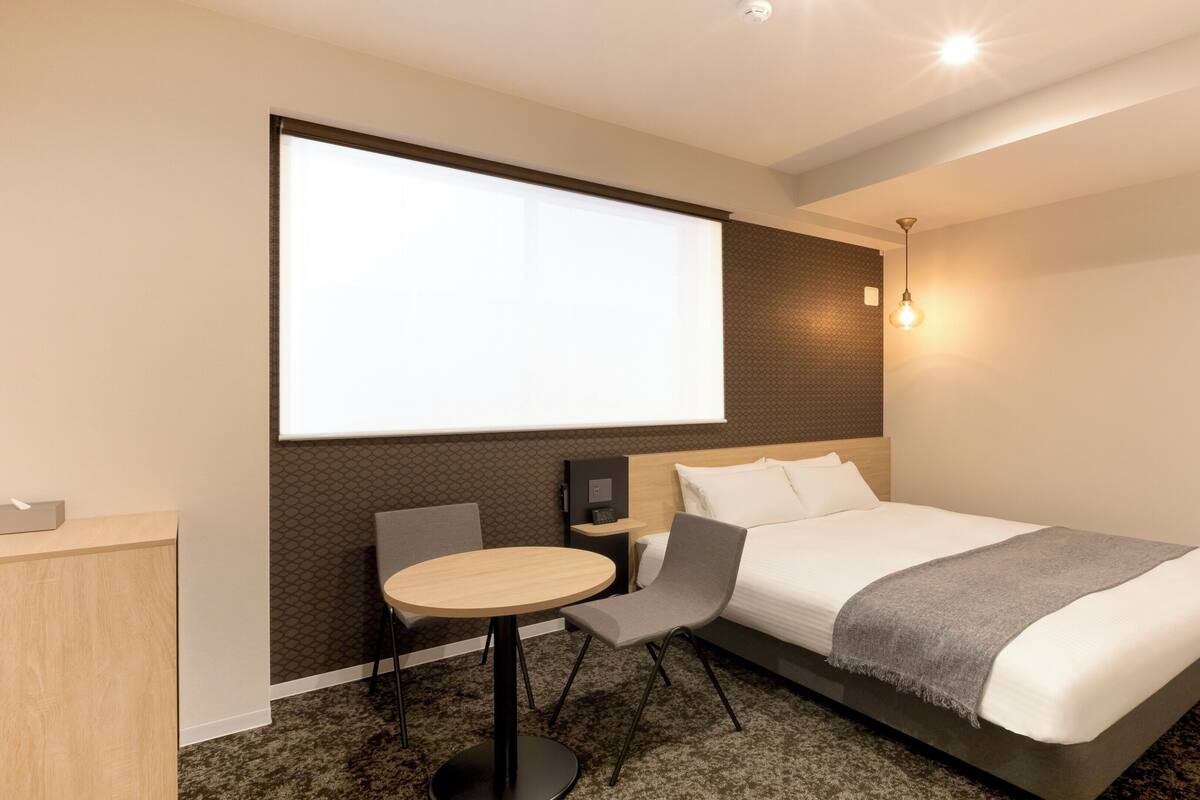 Quarto de hotel moderno com uma cama grande, uma mesa, uma cadeira, tela de projetor e luminária fixada na parede.