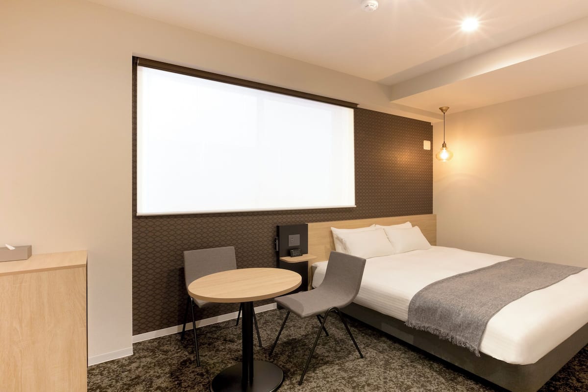 A modern hotel room with a large bed, a desk, a chair, a projector screen, and a wall-mounted light fixture.