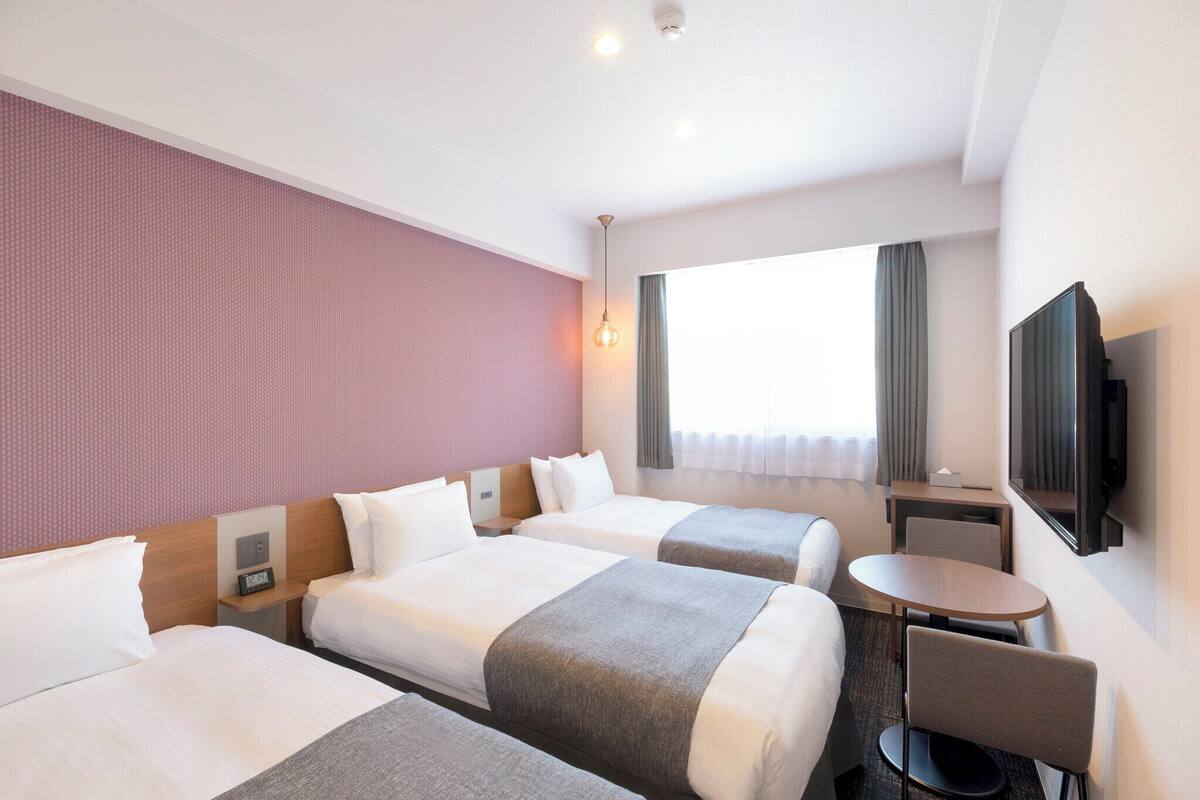 Quarto de hotel com duas camas, uma mesinha, uma cadeira, televisão e janela com cortinas.
