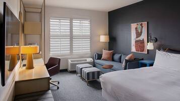 Quarto de hotel com cama, escrivaninha com luminária, sofá, poltrona e uma grande janela com persianas.