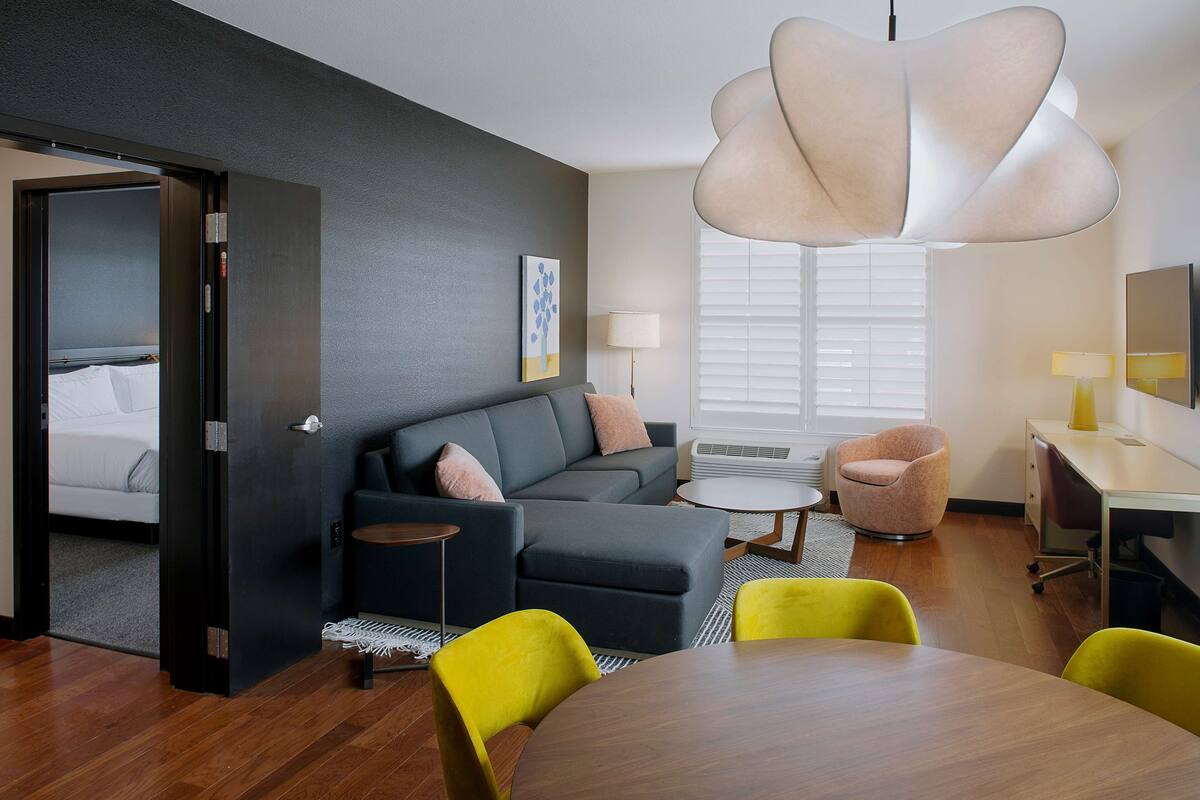Een moderne hotelkamer met een donkergrijze bank, een ronde houten eettafel en een felgele stoel. Er is een groot raam met jaloezieën, een nachtkastje met een lamp en een klein rond tafeltje met een lamp.