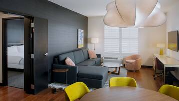 Una habitación de hotel moderna con un sofá gris oscuro, una mesa redonda de madera y una silla amarilla brillante. Hay un ventanal con persianas, una mesita de noche con lámpara y una pequeña mesa redonda con una lámpara.