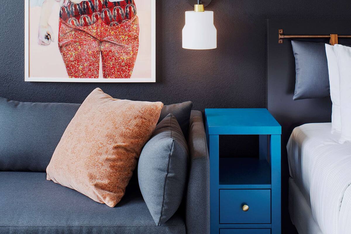 Een moderne slaapkamer met een donkere wand, een lijstwerk met kunst, een blauwe nachtkastje en een grijze bank.