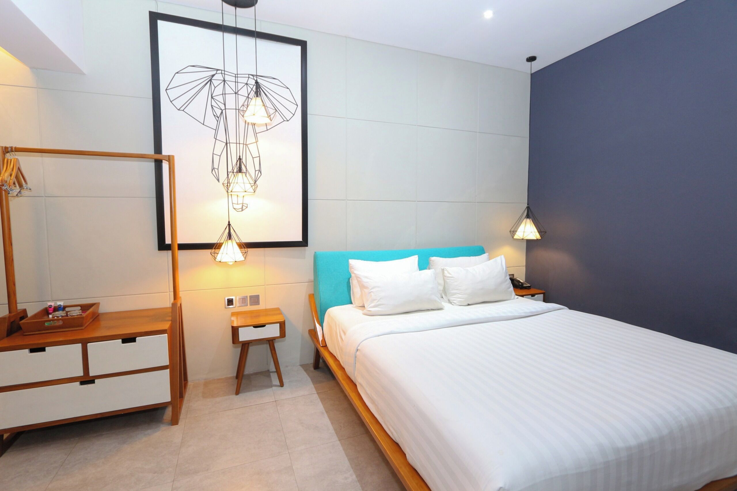 A modern hotel room with a bed, bedside tables, a wooden dresser, and a framed artwork on the wall.