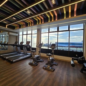 A gym with treadmills, elliptical machines, and a large window with a city view. - Hotel Maximillian (Karimun)