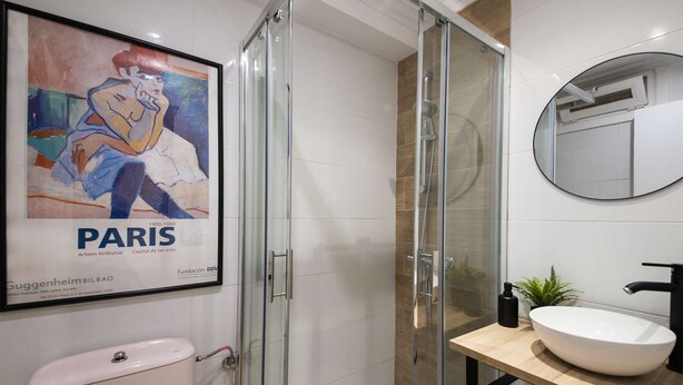 Un baño moderno con ducha acristalada, inodoro blanco, tocador de madera con un lavamanos grande y blanco, y una obra de arte enmarcada de una mujer en la pared.
