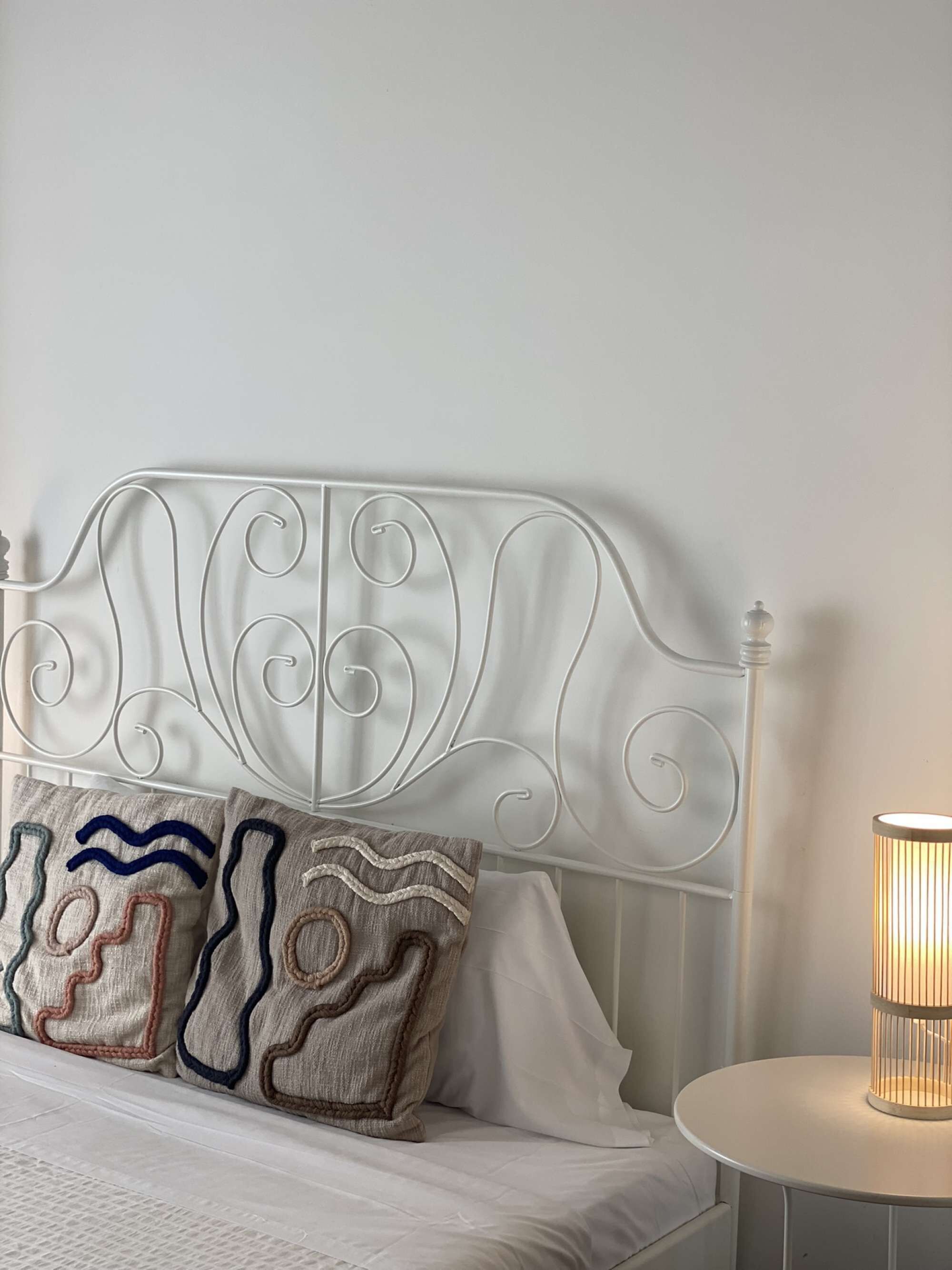 Een bed met een wit metalen hoofdeinde, twee decoratieve kussens en een nachtkastje met een lamp.