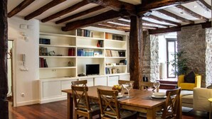 A dining area with a wooden table and chairs, a bookshelf filled with books, and a stone wall in the background. - Spacious, Central and Authentic Family Apartment (Donostia)