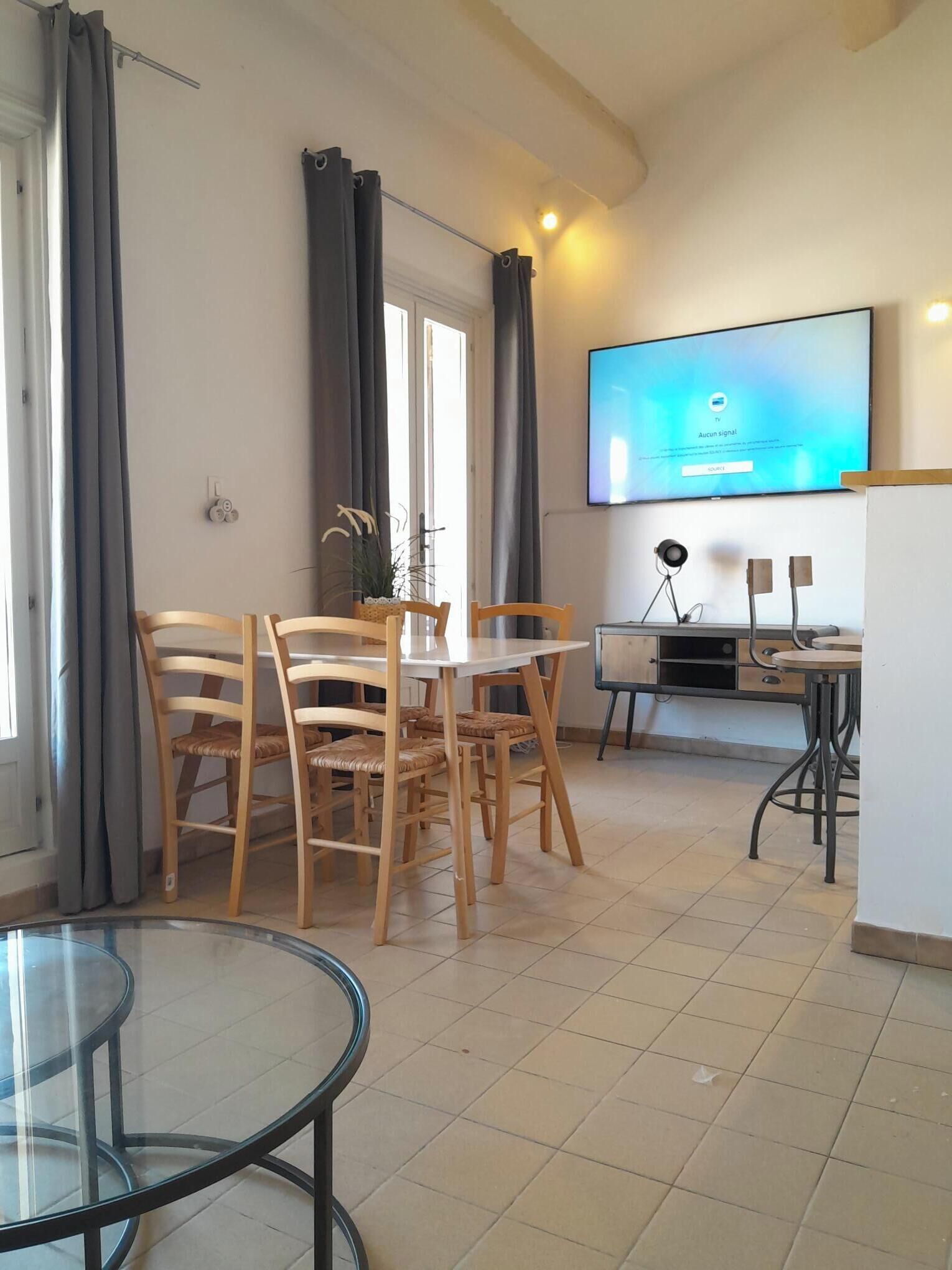 A modern living room with a dining area, a flat-screen TV mounted on the wall, and a small side table with a lamp.