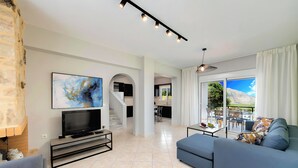 Sala de estar moderna com televisão de tela plana, sofá azul e área de jantar com vista para o exterior.