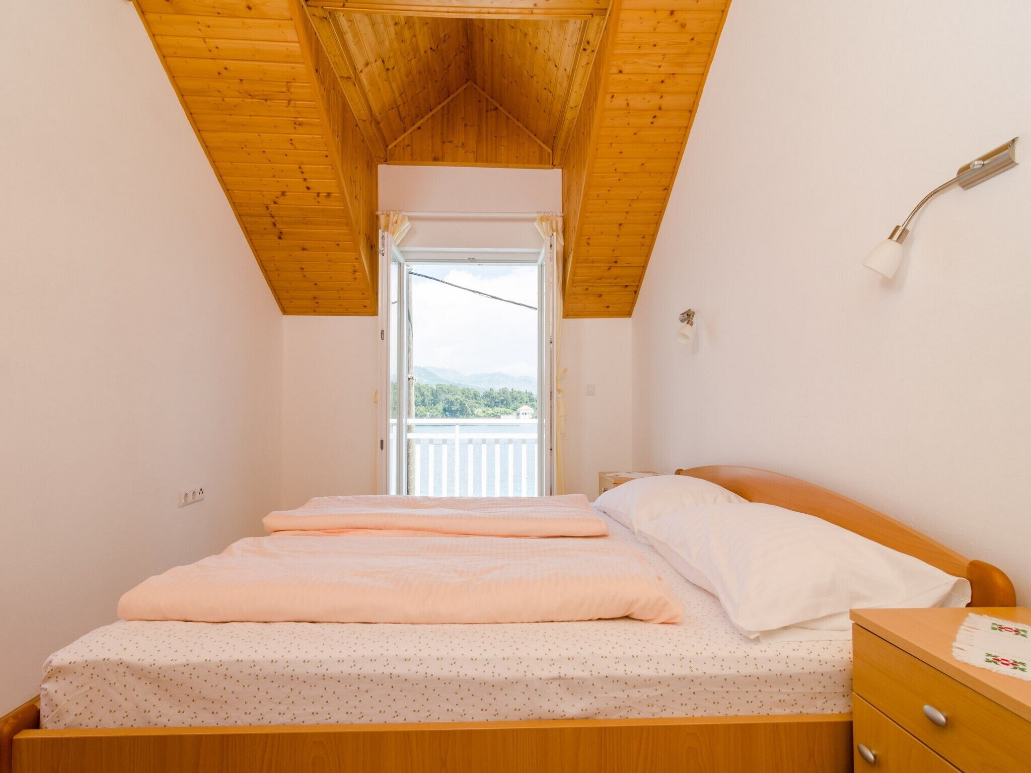 Ein Schlafzimmer mit Holzboden, einem Bett mit rosa und weißen Bettwaren, einem Nachttisch und Blick auf Grün hinter einer geöffneten Tür.