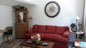 A living room with a red sofa, a wooden cabinet, a round wall clock, and a small table with a vase of flowers.
