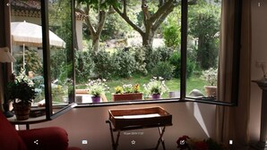 A room with a large window overlooking a garden with various potted plants and a wooden bench. - Charming and quiet cottage (Aix-en-Provence)