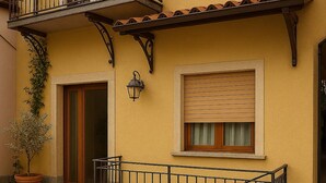 A yellow building with a wooden door, a window with a wooden blind, a potted plant, and a metal railing.