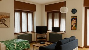 A modern living room with a sofa, a TV, a small table, and a bed with a green leaf-patterned bedspread.