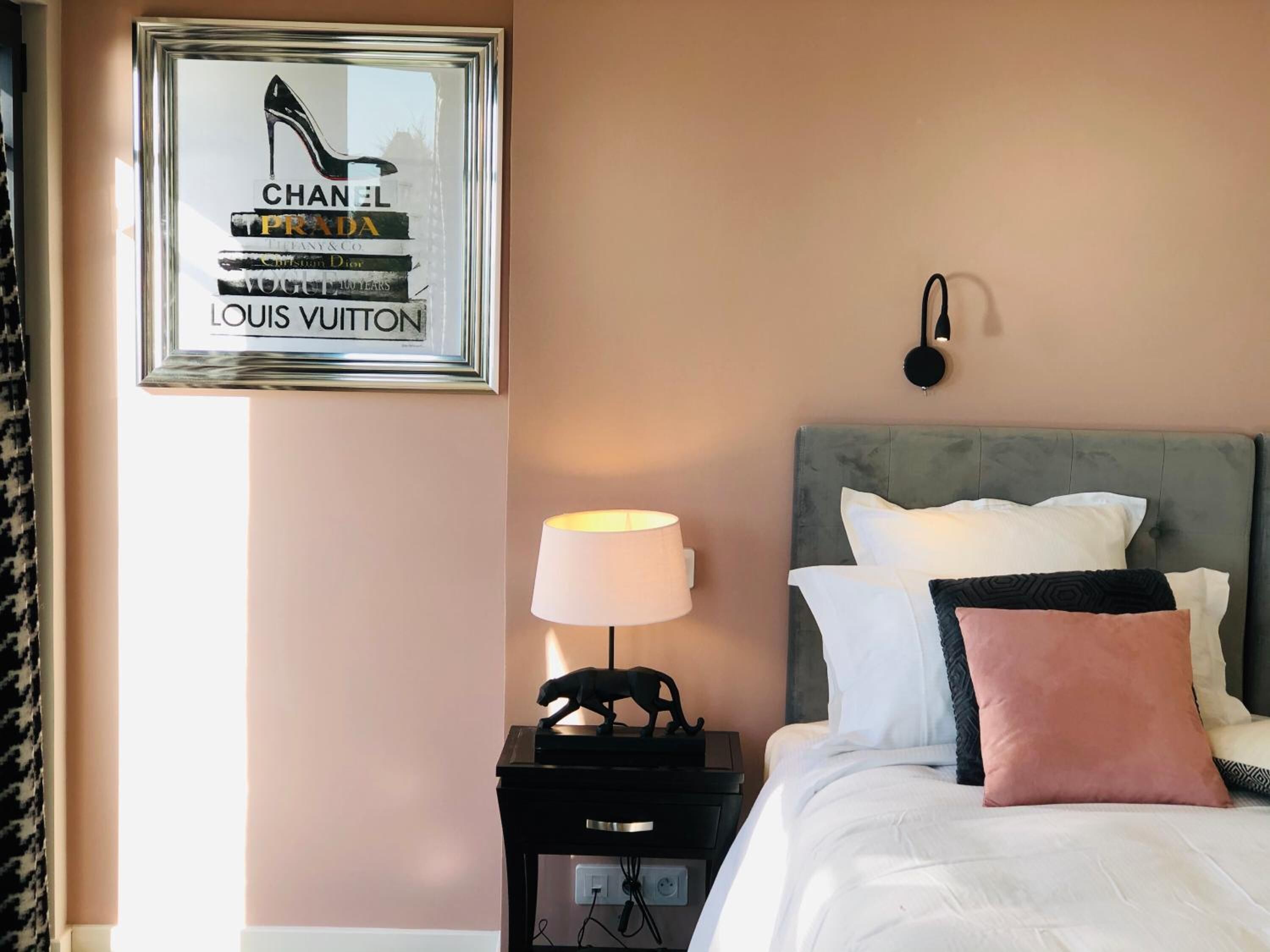 A hotel room with a bed, a nightstand with a lamp, a framed poster of luxury brands, and a wall-mounted light fixture.