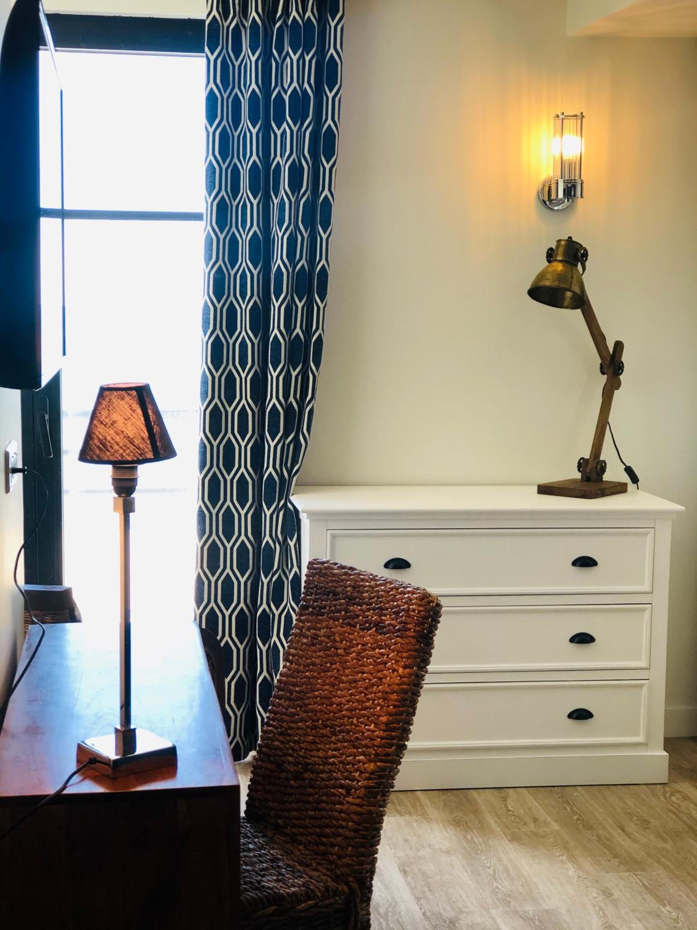 A room with a white dresser, a lamp, a wicker chair, and a patterned curtain.