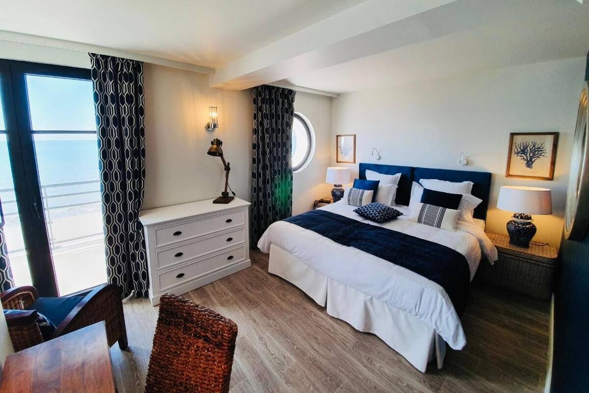 A modern bedroom with a large bed, bedside tables, a desk, and a view of the sea.