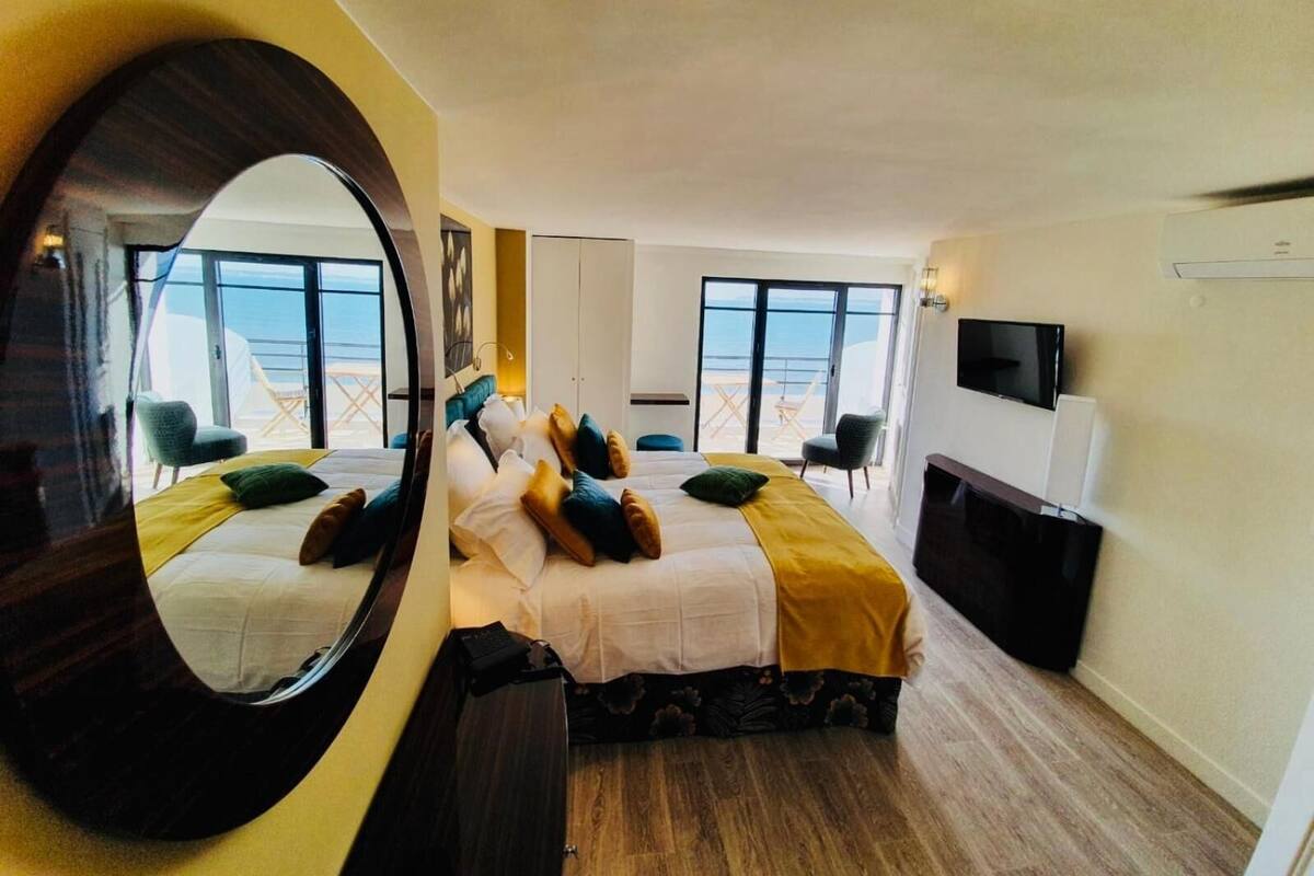 A hotel room with a bed, a TV, a desk, and a large window with a view of the sea.