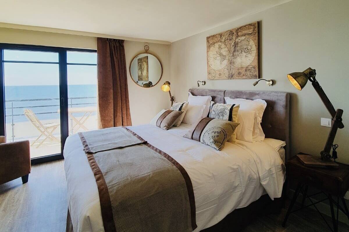 A bedroom with a large bed, a round mirror, a map artwork, a desk with a lamp, and a view of the beach.