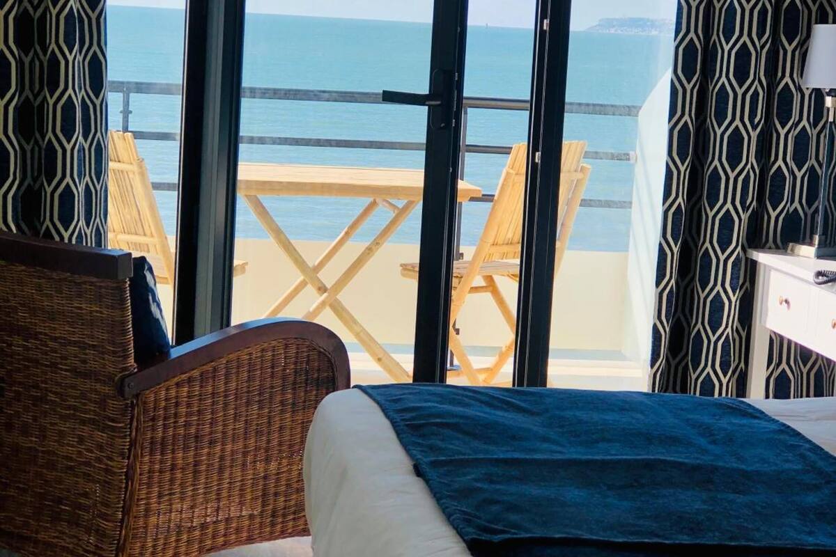 A room with a bed, a wicker chair, and a view of the beach through large glass doors.
