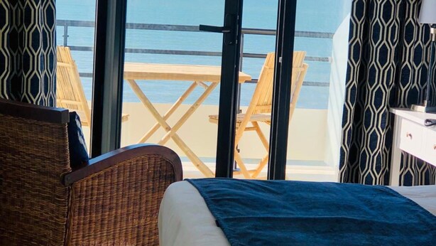 Une chambre avec un lit, un fauteuil en osier et une vue sur la plage, visible à travers de grandes portes vitrées.