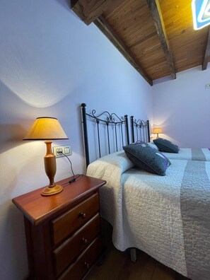 Quarto com mesa de cabeceira de madeira, luminária, cama com duas almofadas e teto de madeira.