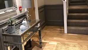 Una escalera con peldaños oscuros y balaustres blancos, un espejo con un marco ornamentado, una puerta de madera y una pequeña mesa con una lámpara y objetos decorativos.