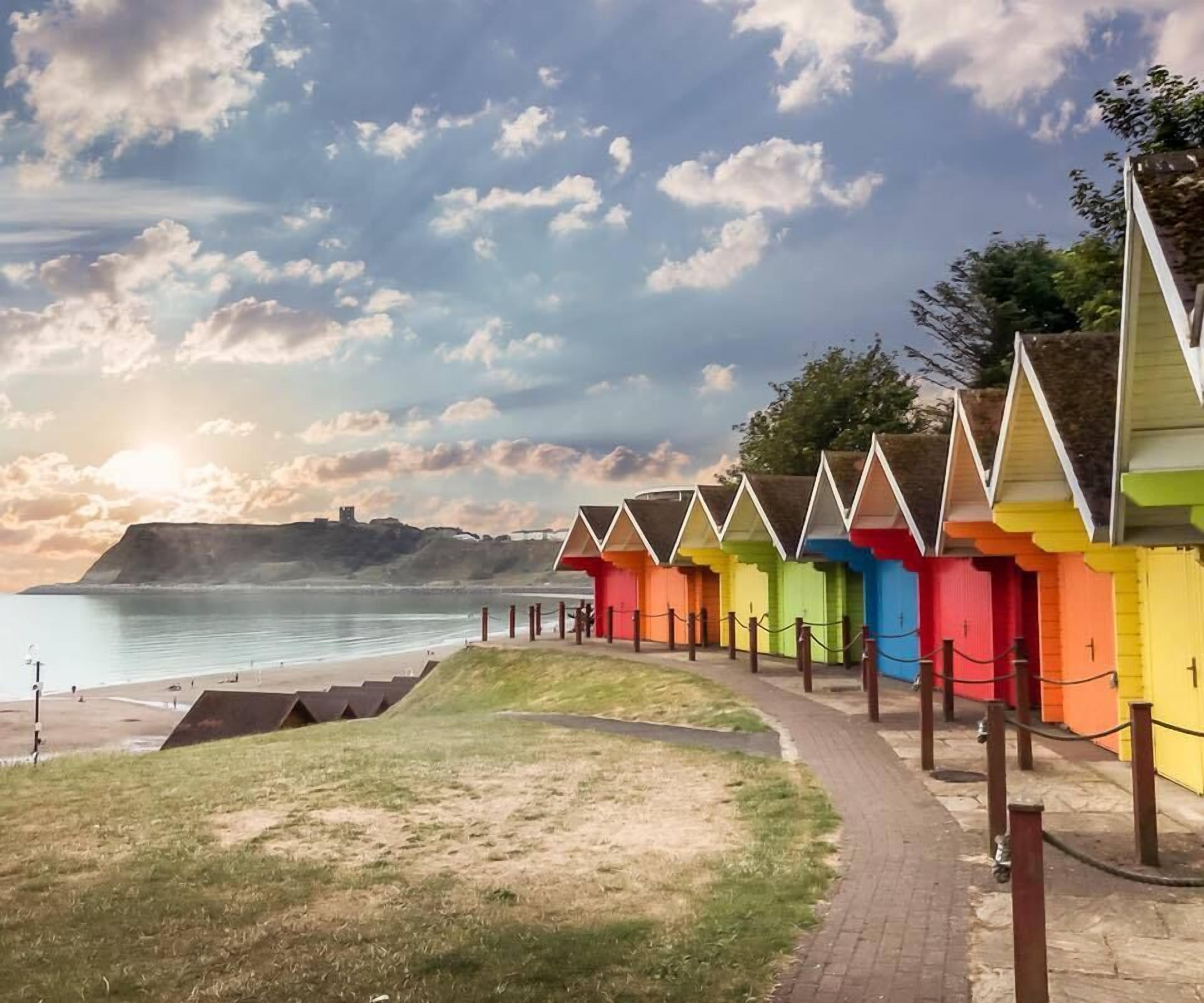 Fila de cabañas de playa coloridas a lo largo de un paseo marítimo con vista despejada al océano y una colina distante.