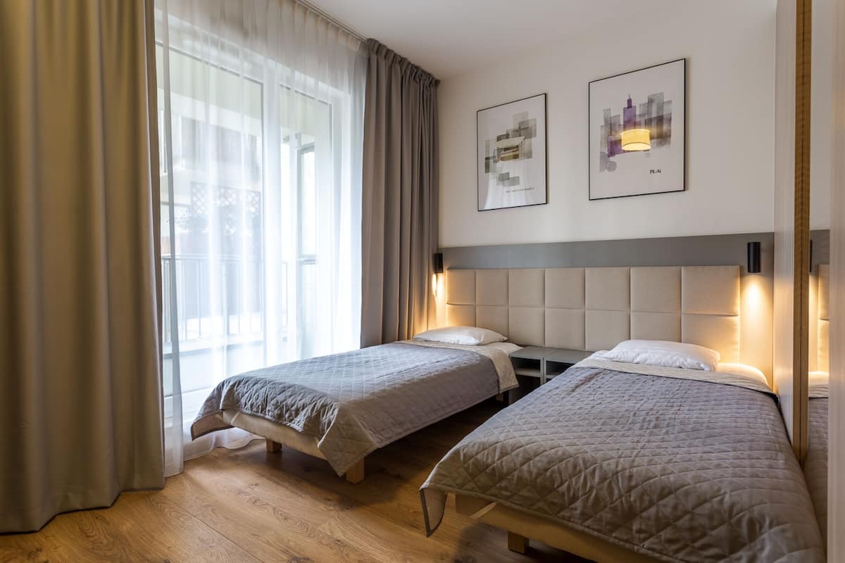 A hotel room with two beds, a large window with sheer curtains, and two framed pictures on the wall.