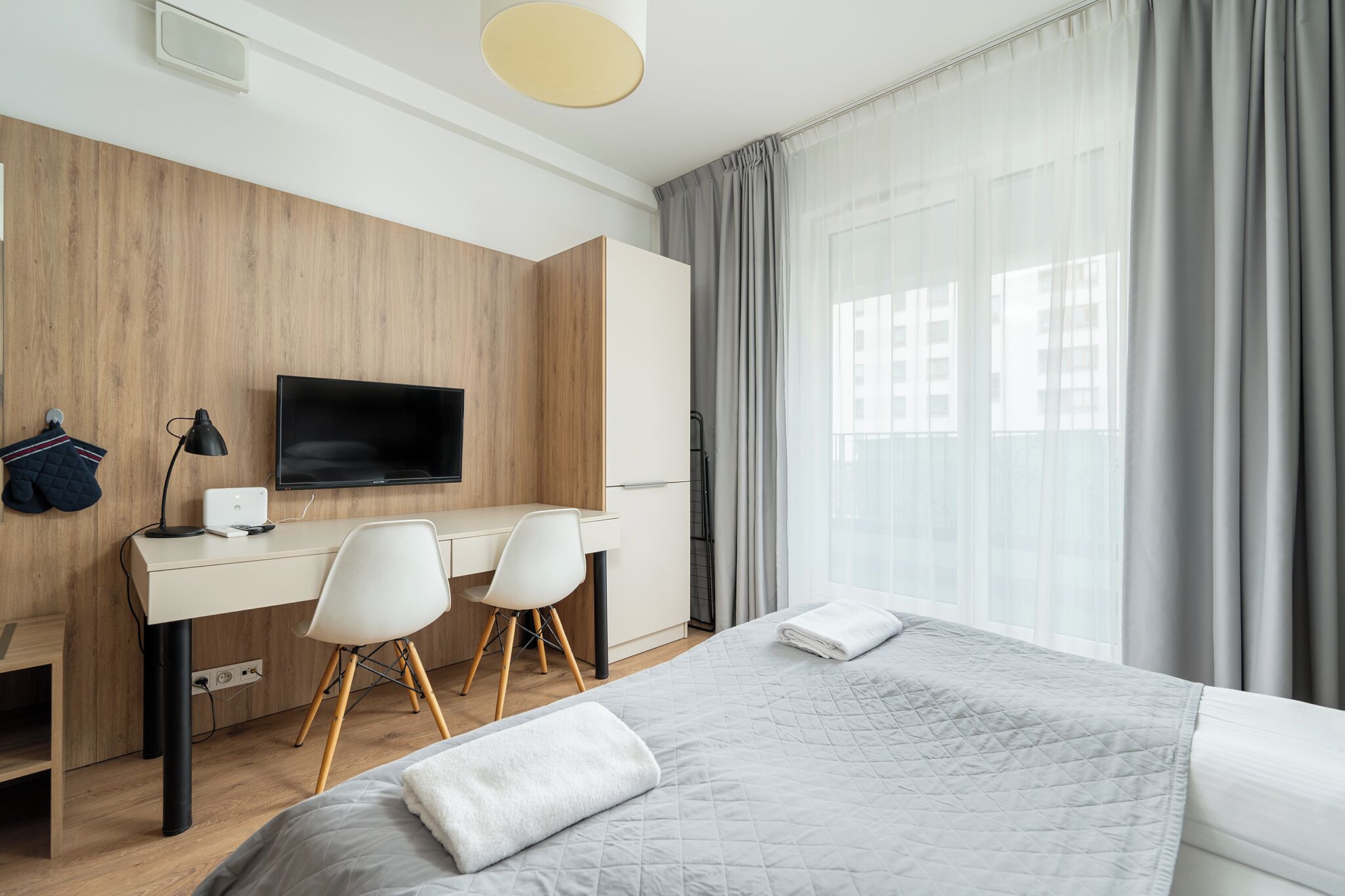 Une chambre d’hôtel moderne équipée d’un lit, d’un bureau avec deux chaises, d’une télévision et d’une grande fenêtre avec des rideaux.