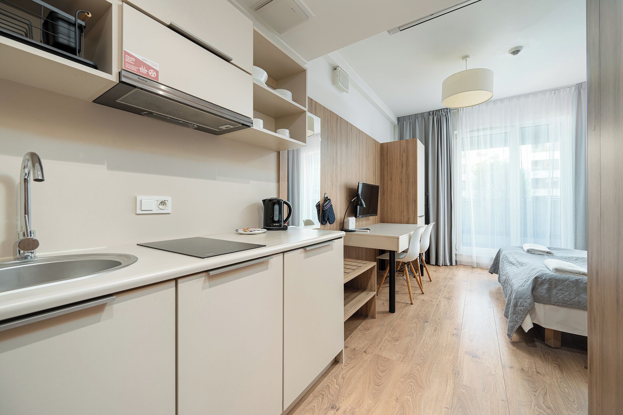 Cocina moderna con lavamanos, estufa y encimera. Hay un comedor con mesa y sillas, y una cama con colcha gris.