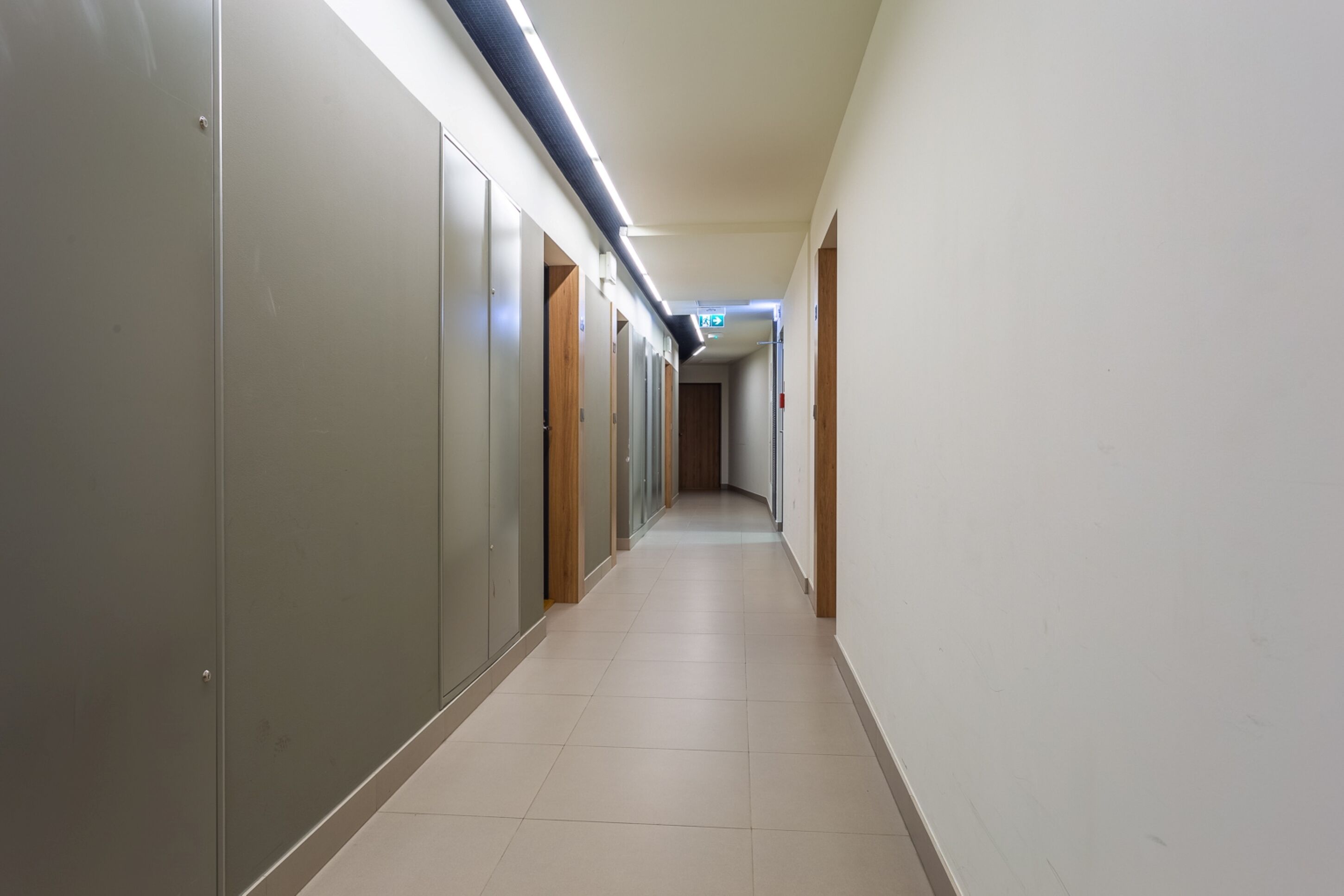 A long, narrow hallway with lockers on one side and doors on the other, leading to a room at the end.