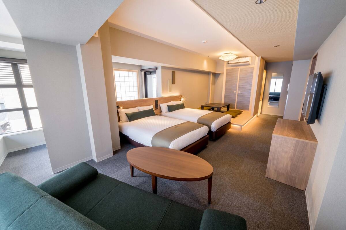 A hotel room with two beds, a sofa, a wooden table, and a TV mounted on the wall.