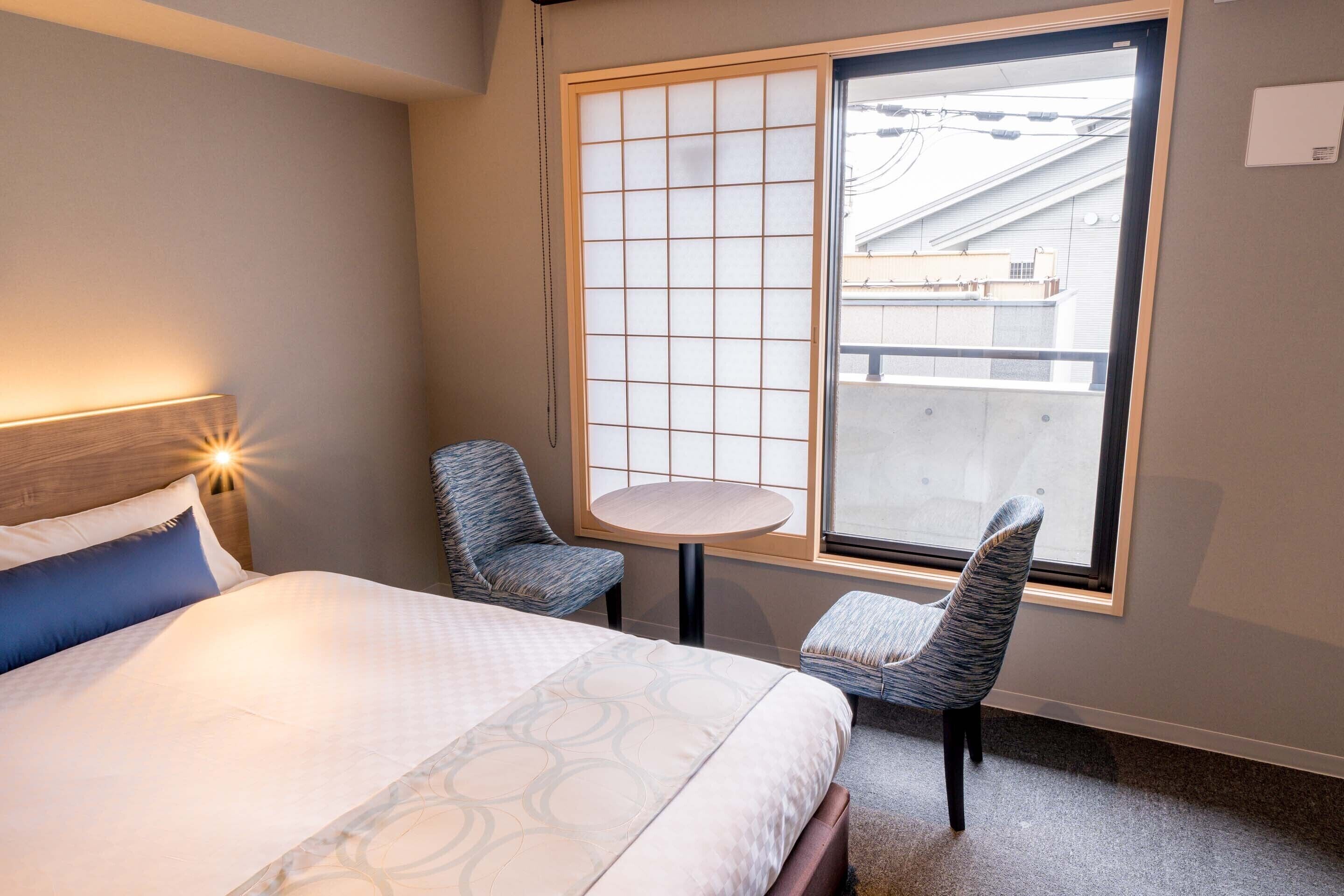 Habitación de hotel con cama, una mesita redonda pequeña, dos sillas y un ventanal con vistas a un edificio.
