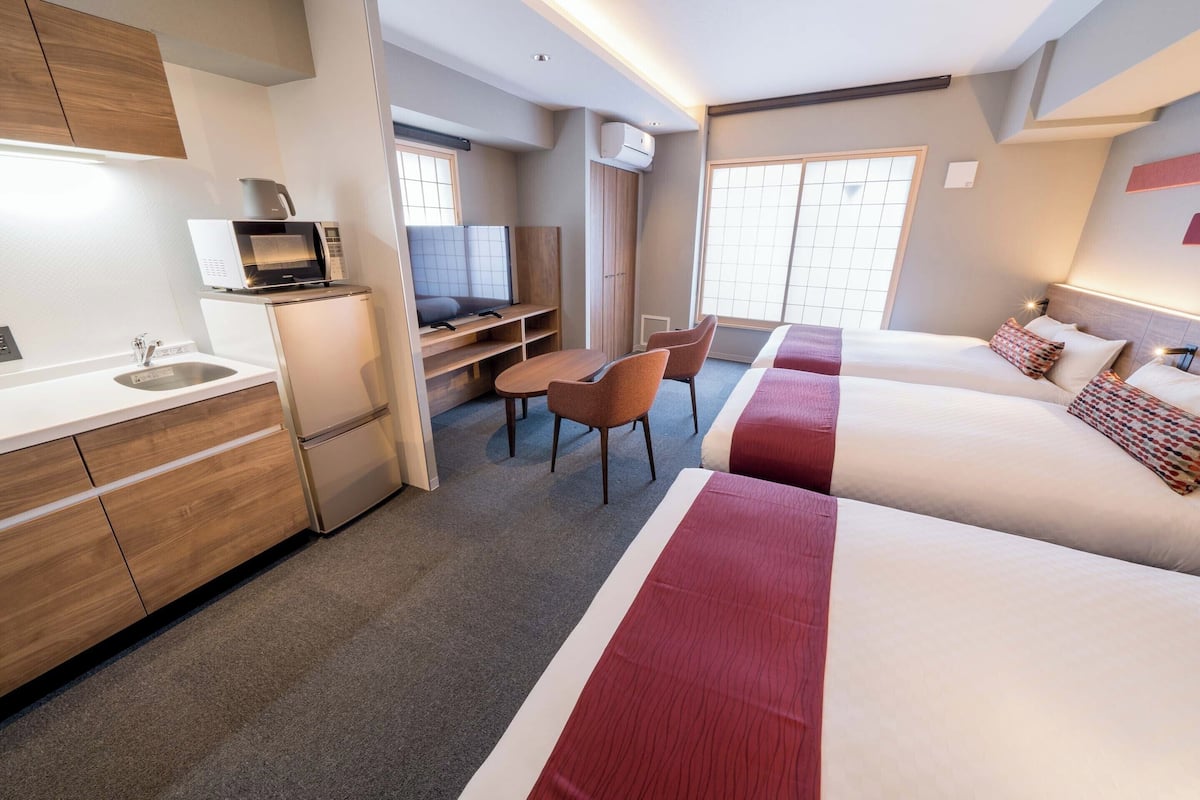 A hotel room with a kitchenette, a TV, a desk with chairs, and a bed with red bedding.