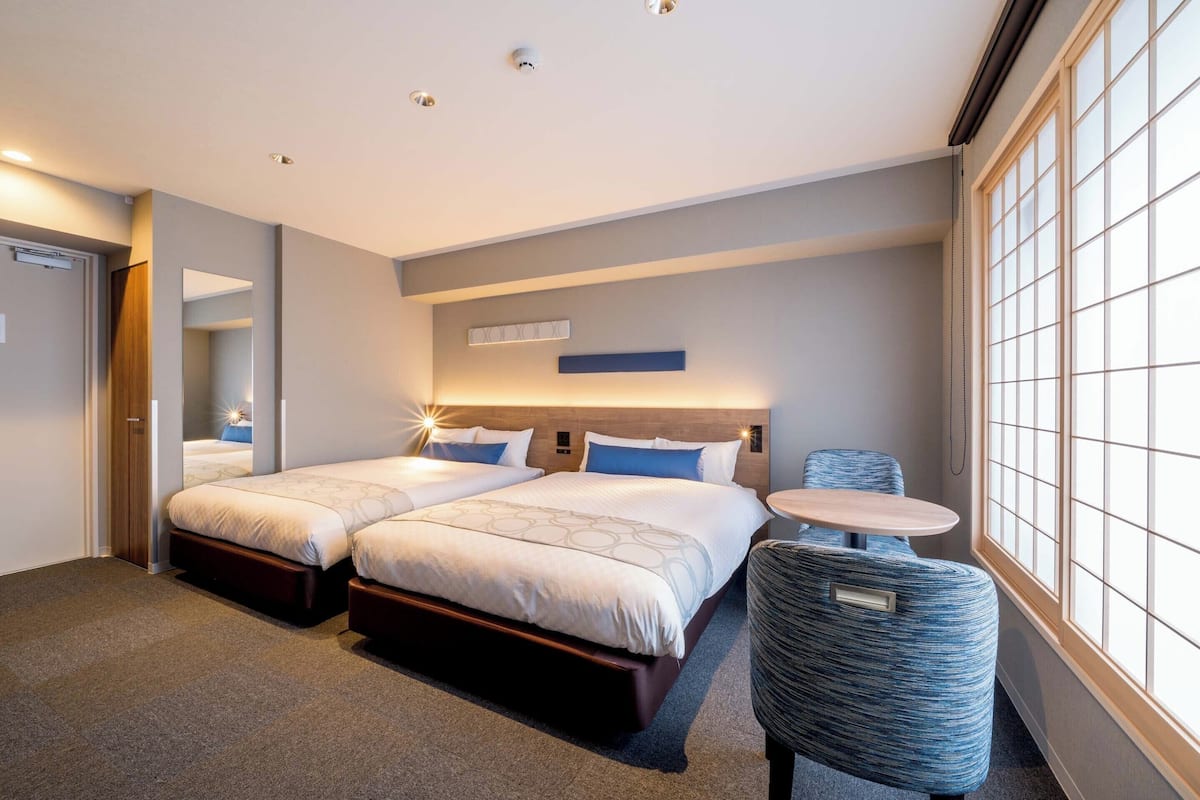 A hotel room with two beds, a small round table, a chair, and a large window.