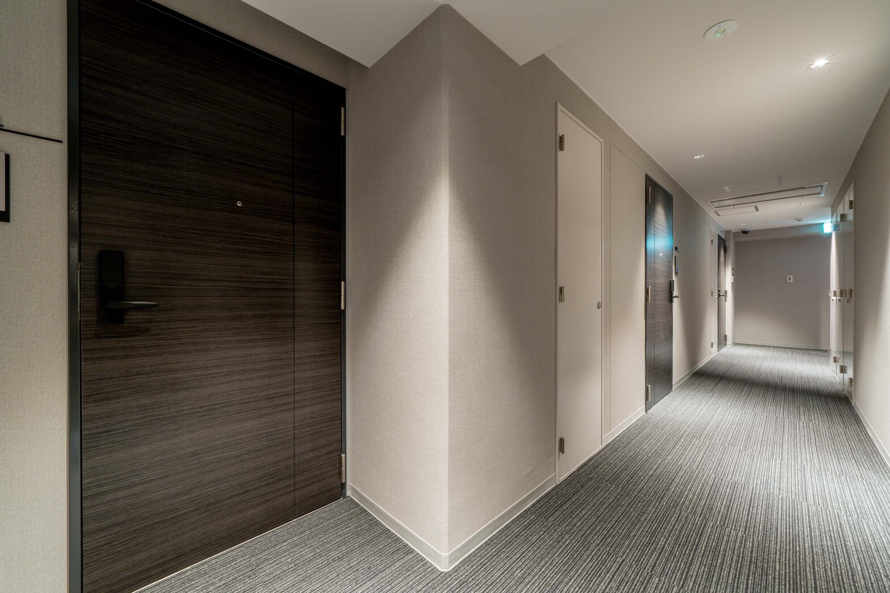 A modern hotel corridor with a dark wood door, a light-colored wall, and a carpeted floor.