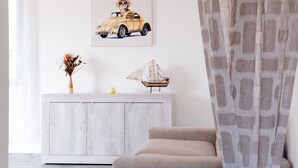 Ett vardagsrum med ett vitt skåp, en beige soffa, en tavla på väggen som föreställer en hund i en bil, en vas med torkade växter och en modell av ett skepp.