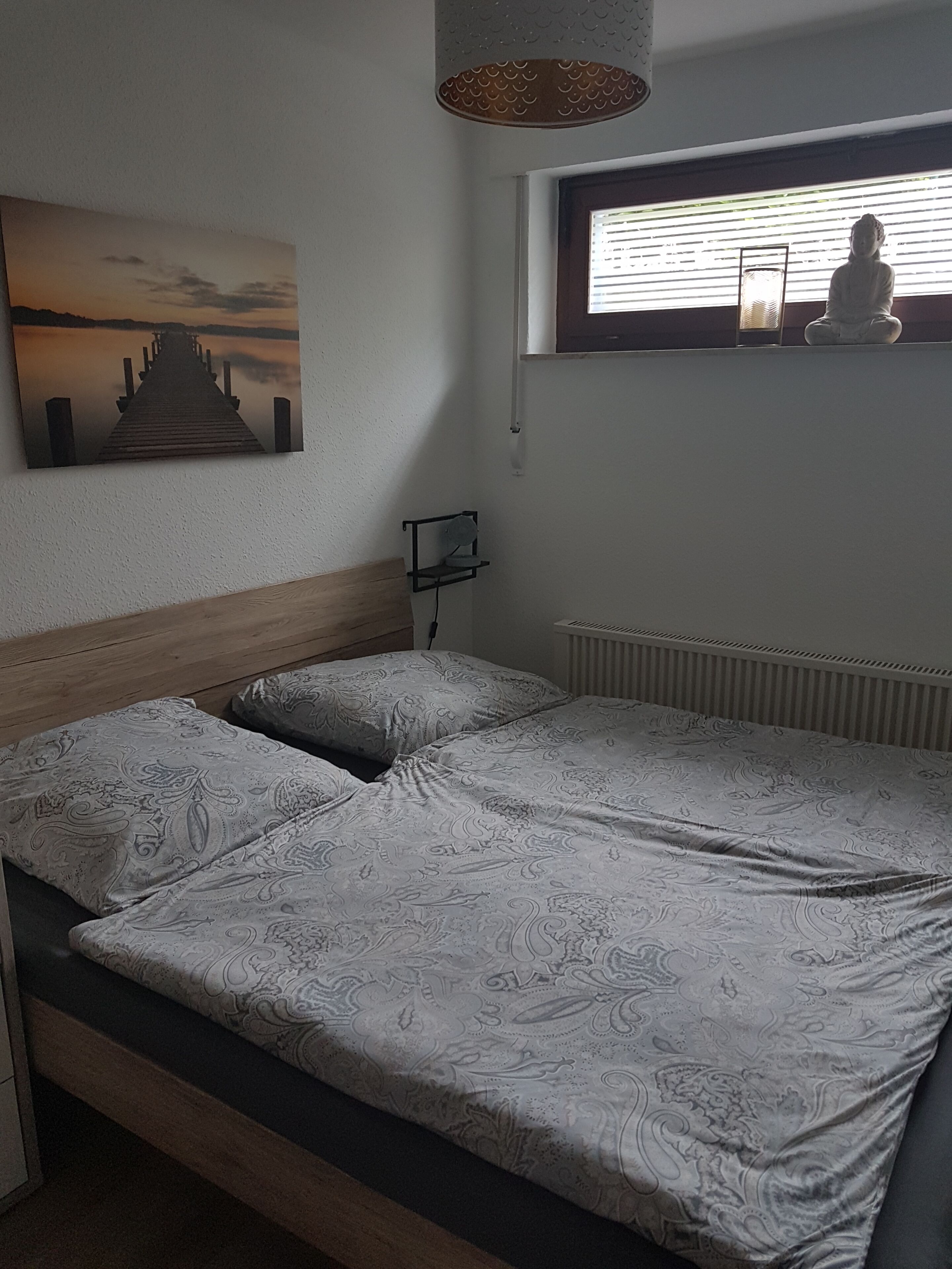 Un dormitorio con cabecera de madera, cama con ropa de cama estampada, estante con lámpara, ventana con persianas y un cuadro de un muelle.