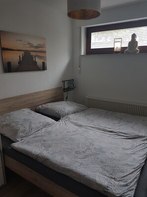 Une chambre avec une tête de lit en bois, un lit recouvert d’une literie à motifs, une étagère fixée au mur avec une lampe, une fenêtre avec des stores et un tableau représentant une jetée.